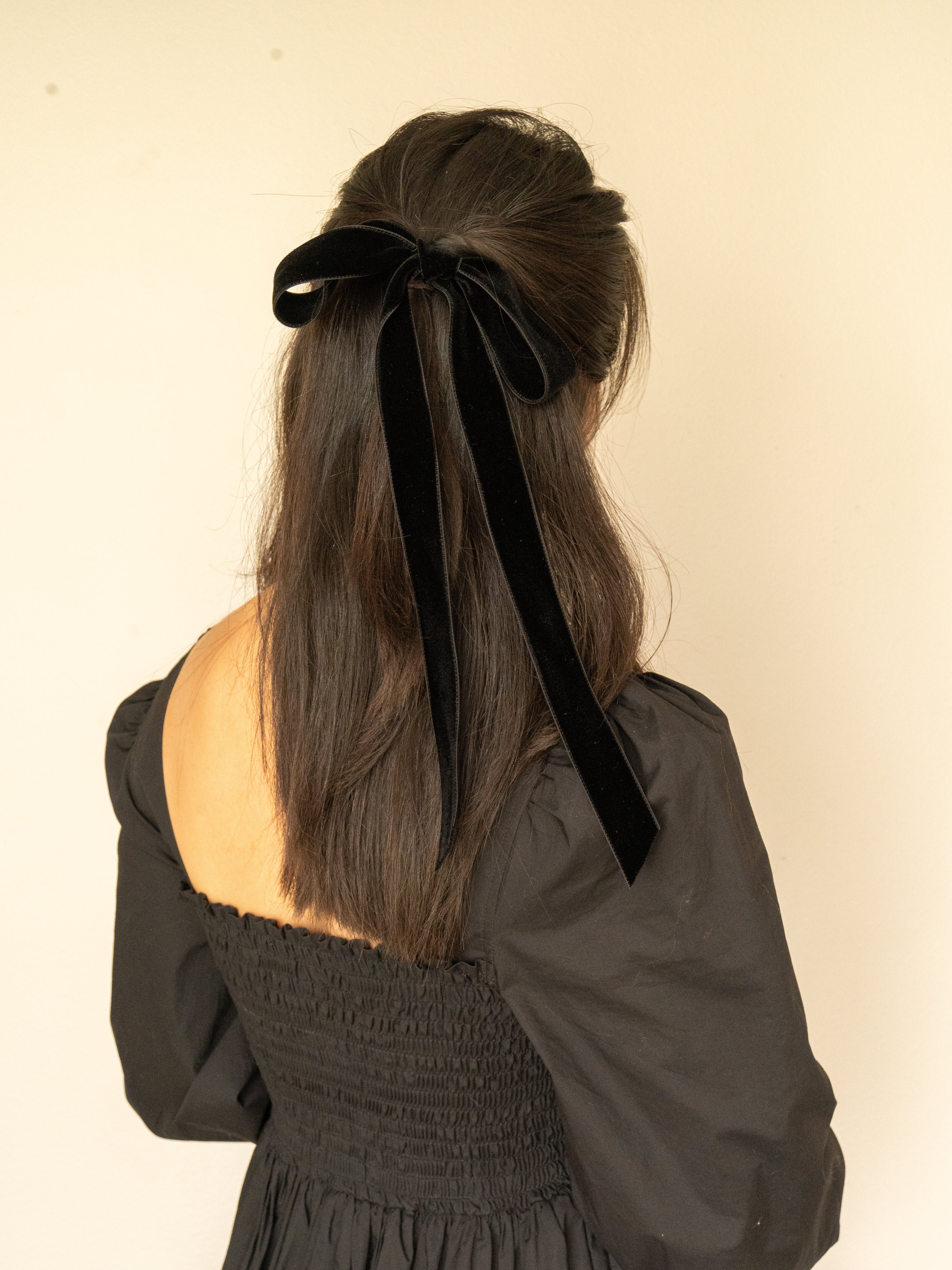 Person wearing a black dress with a large black velvet bow in their hair against a plain background